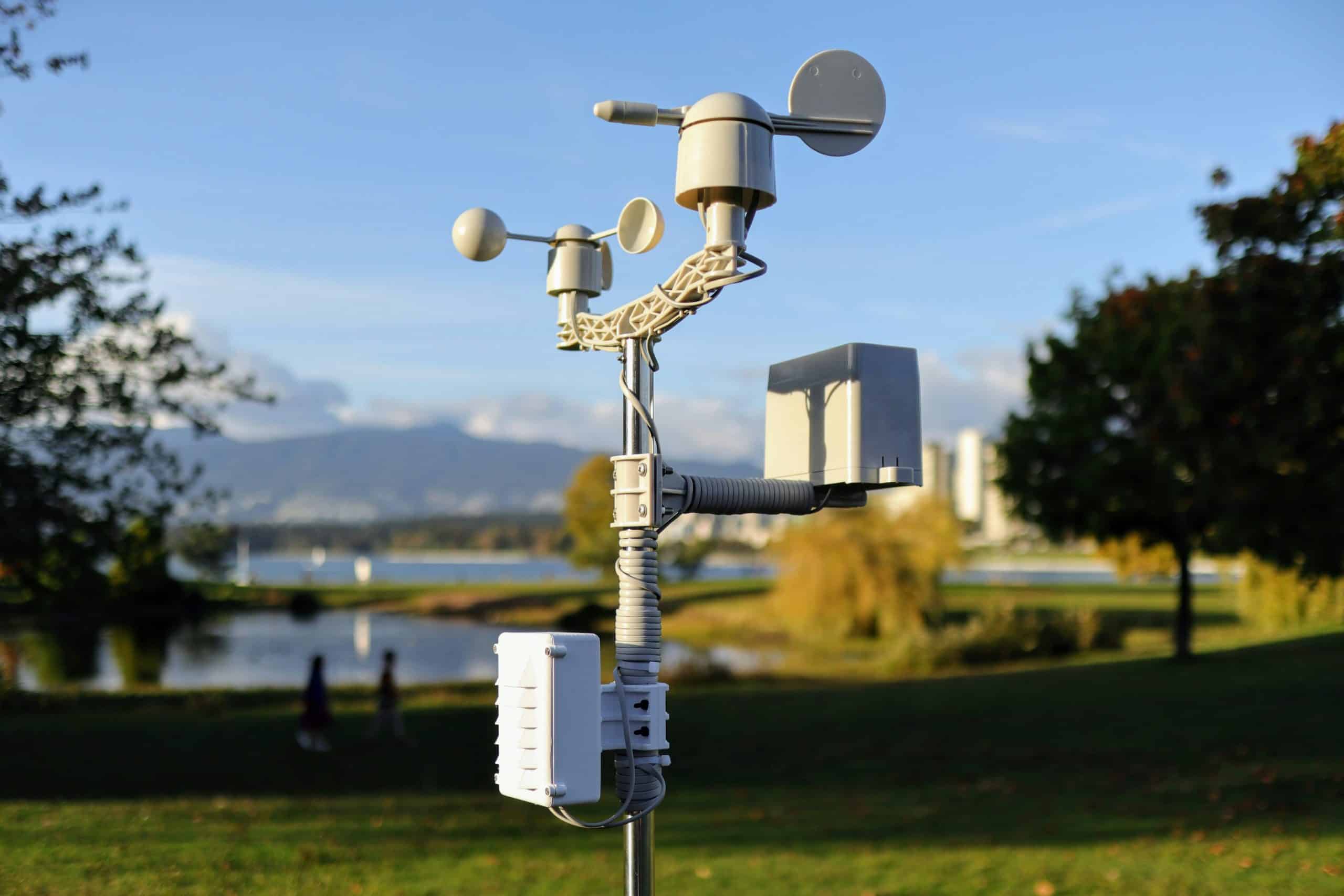 Close-up of an outdoor weather and environmental sensor station equipped with anemometers and other measurement devices, set against a blurred background of a park, water, and distant mountains.